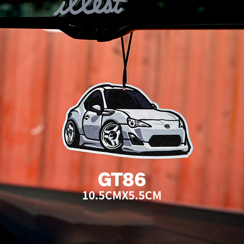 Car air freshener shaped like a GT86 car hanging from a rearview mirror with an orange wall background.
