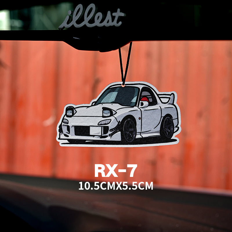 Car air freshener with RX-7 design hanging from a rearview mirror against an orange wall.