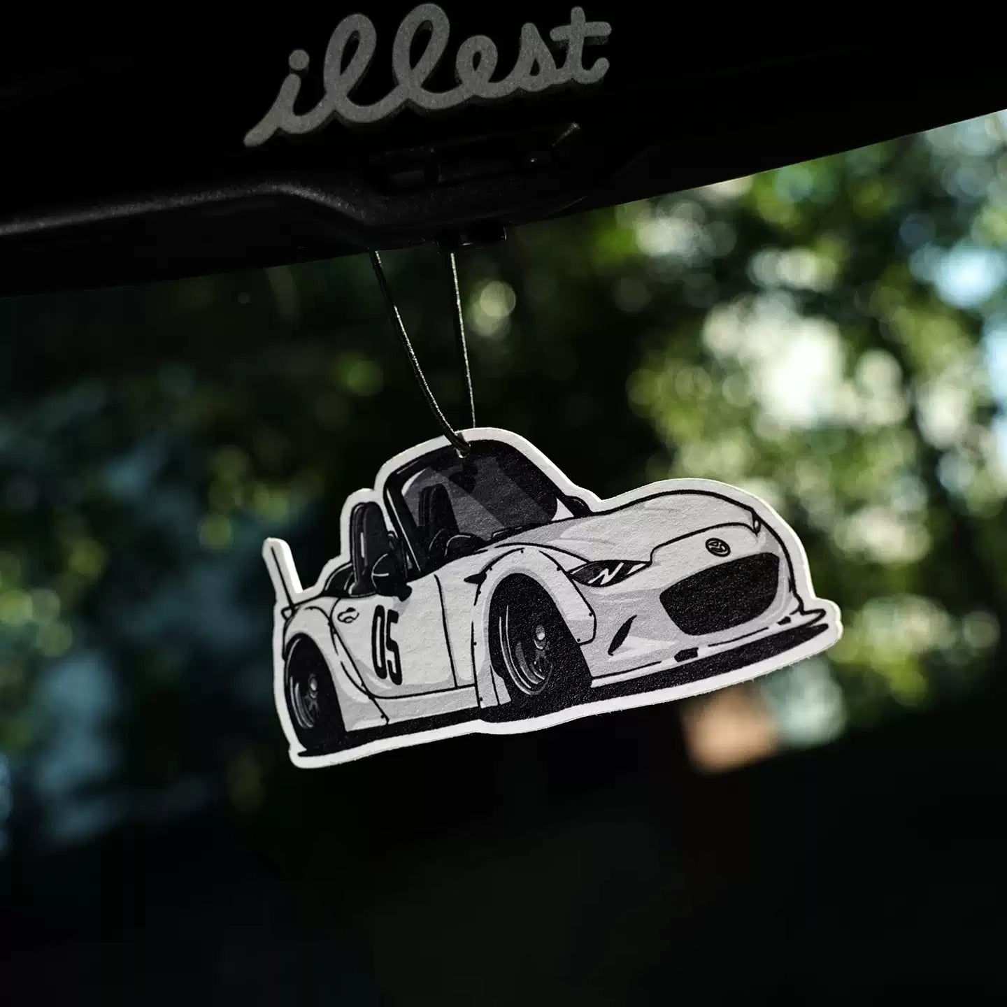 Car air freshener shaped like a white sports car with 'illest' branding on a blurred green background.