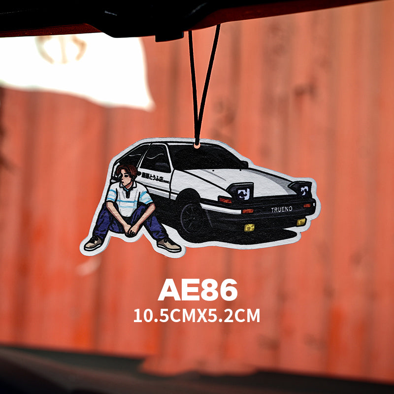 Car air freshener with a design of a person sitting next to a white Toyota AE86 on an orange background.