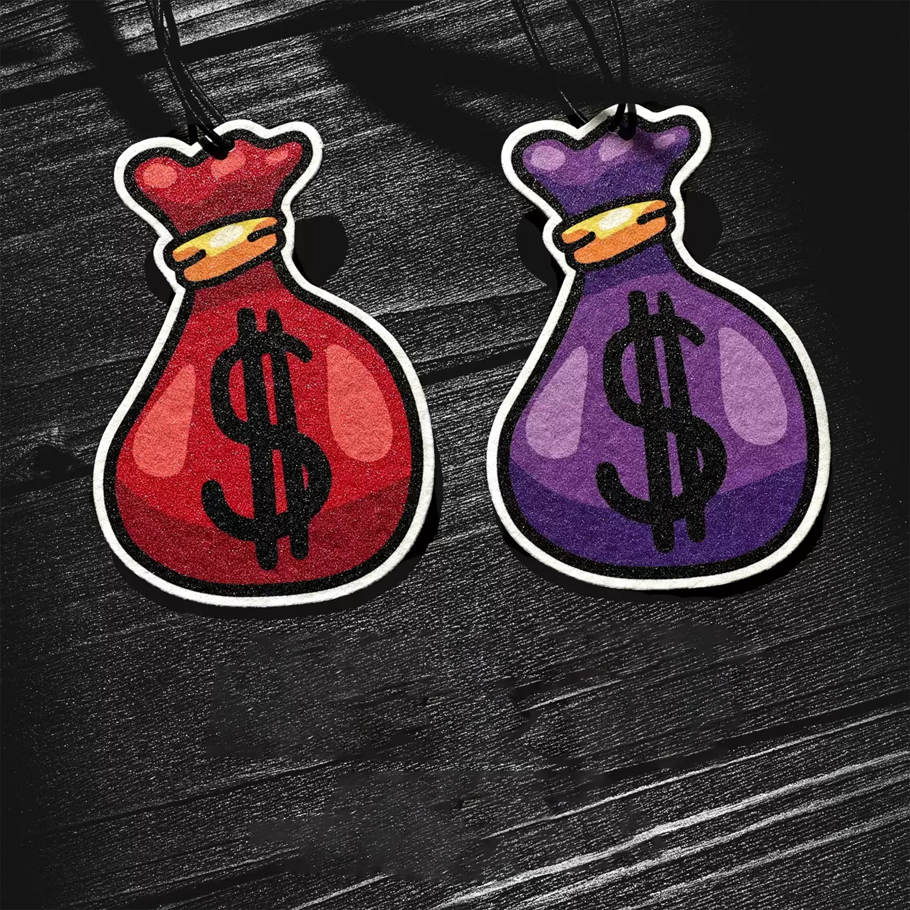 Two bag-shaped air fresheners, one red and one purple, with dollar sign symbols on a dark background.