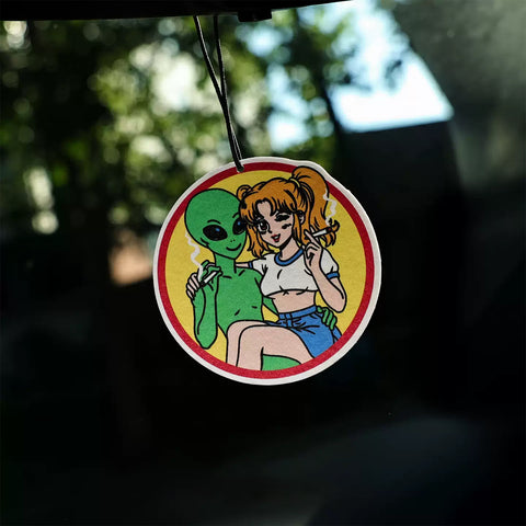 Circular air freshener with an alien and a person design, hanging against a blurred natural background.