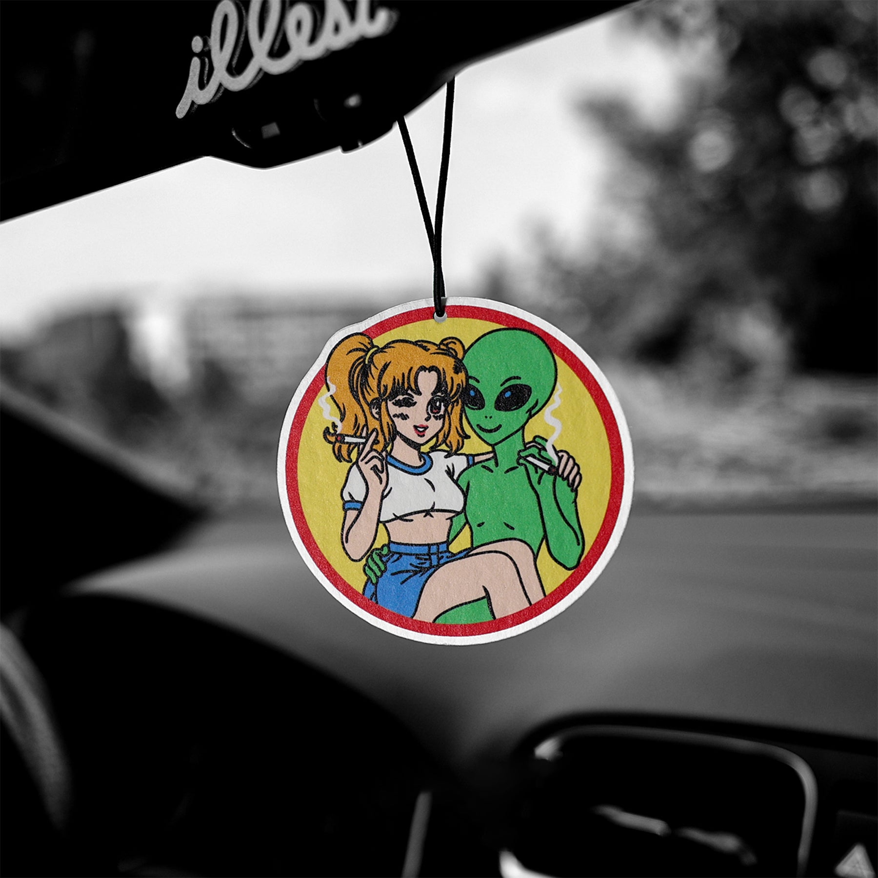 Car air freshener with cartoon design of a woman and alien on a yellow background, hanging from a car's rearview mirror.