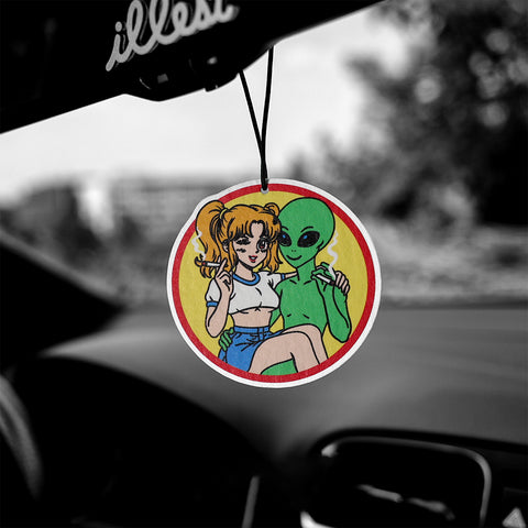 Car air freshener with cartoon design of a woman and alien on a yellow background, hanging from a car's rearview mirror.