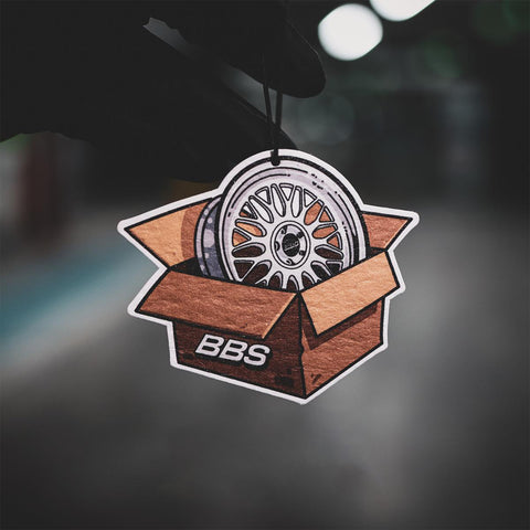 Car air freshener shaped like a wheel in a box with 'BBS' branding, hanging against a blurred background.