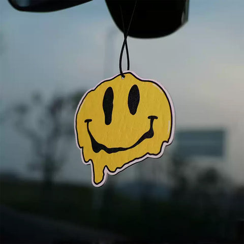Yellow smiley face air freshener hanging from a car mirror with a blurred outdoor background.