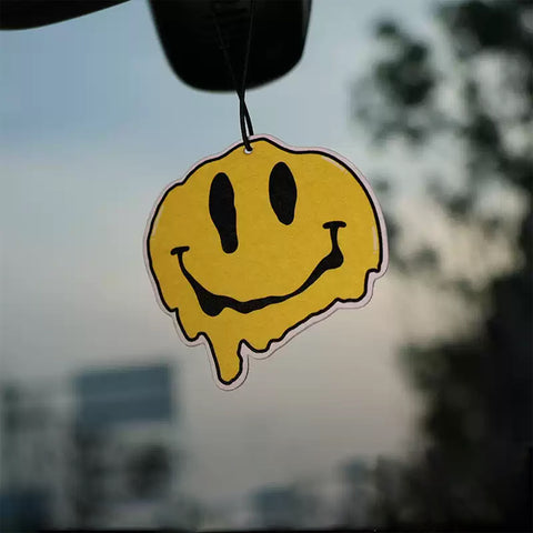 Smiley face air freshener hanging from a car mirror with a blurred outdoor background